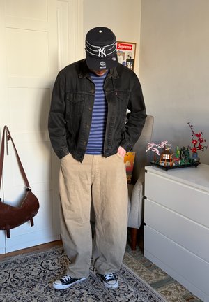 Persona con chaqueta de mezclilla negra, suéter azul a rayas, pantalones beige, gorra negra de NY y zapatillas negras, de pie en interiores cerca de una cómoda blanca y una silla.