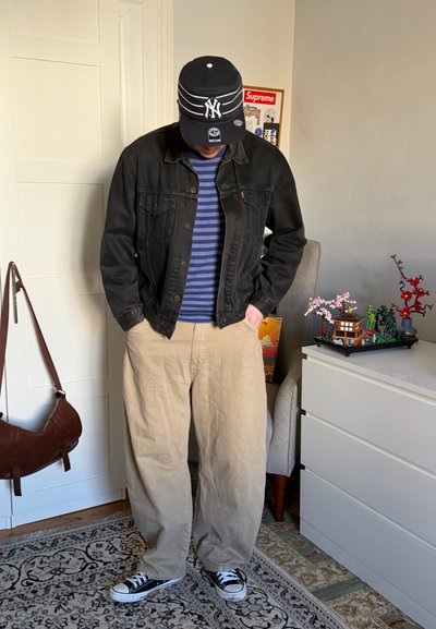 Persona con chaqueta de mezclilla negra, suéter azul a rayas, pantalones beige, gorra negra de NY y zapatillas negras, de pie en interiores cerca de una cómoda blanca y una silla.