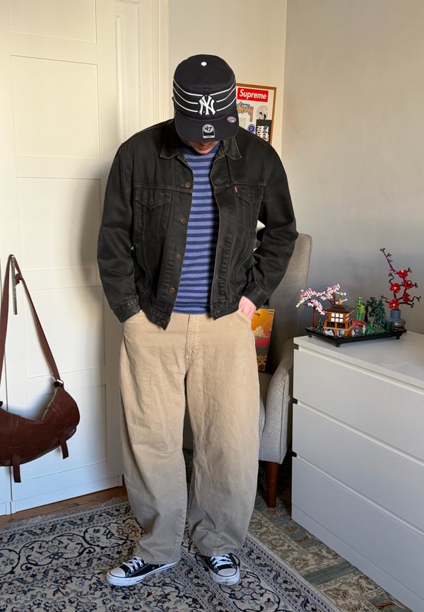 Person som har på seg svart denimjakke, blå stripete genser, beige bukser, svart NY-cap og svarte joggesko som står innendørs ved siden av en hvit kommode og stol.