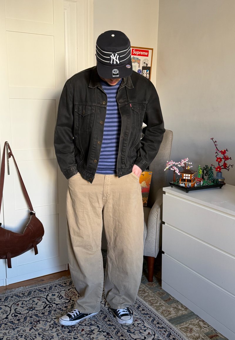 Persona con chaqueta de mezclilla negra, suéter azul a rayas, pantalones beige, gorra negra de NY y zapatillas negras, de pie en interiores cerca de una cómoda blanca y una silla.