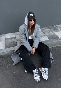 Manteau oversized gris avec une capuche, t-shirt blanc, pantalon de jogging noir et baskets grises. Assis contre un mur gris sur un pavement texturé.