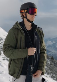 Skiklær med en svart hjelm med oransje detaljer, speilede goggles, en olivenfarget jakke og en svart skjorte, mot en snødekt bakgrunn.