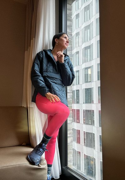 Chaqueta acolchada negra, leggings rosas, zapatillas grises con calcetines con estampado. De pie junto a una ventana con arquitectura moderna afuera.