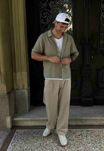 Camisa de cuadros verde claro de manga corta sobre una camiseta blanca, combinada con pantalones beige y zapatillas blancas, de pie frente a puertas oscuras.