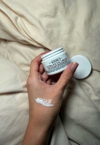 Hånd holder et åbent glas Kiehl's Ultra Facial Cream med creme påført bagsiden af hånden, der hviler på beige stof.