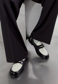 Black leather shoes with white, textured toe caps and a smooth finish. Featuring a decorative strap and a block heel. Soft fabric socks are visible.