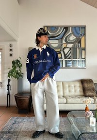 Polo bleu à manches longues avec col blanc, arborant un logo et le numéro 3. Pantalon de survêtement gris et chaussures noires, debout dans un intérieur moderne.