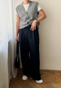 Grå strikket ermeløs genser med frontknapper over en hvit t-skjorte, kombinert med løse, mørke bukser og hvite sneakers. Svart veske inkludert.