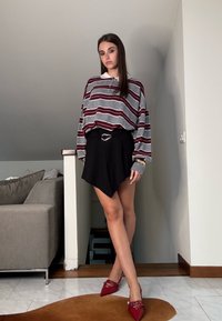 Ravno prugasta siva in burgundska oversize srajca, črna asimetrična krilo z pasom in rdeče špičaste čevlje. Prostroom ima svetle stene.