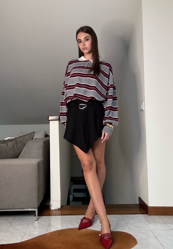 Chemise oversized rayée grise et bordeaux, jupe asymétrique noire avec ceinture, et chaussures pointues rouges. La pièce possède des murs de couleur claire.