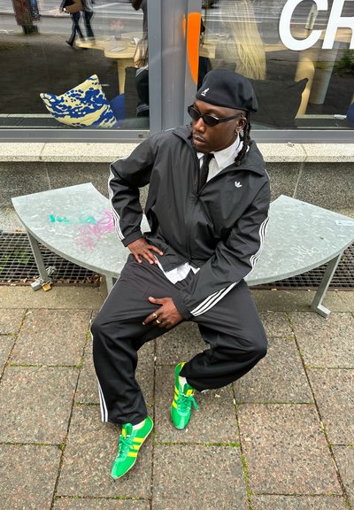 Chándal negro de Adidas con franjas blancas, camisa blanca, corbata negra y zapatillas Adidas verde brillante con detalles amarillos.