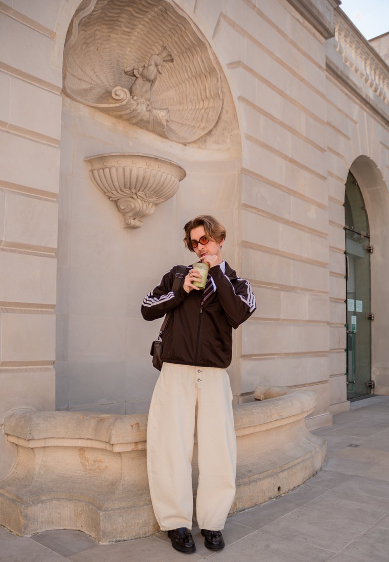 Junge Person mit Sonnenbrille, schwarzer Jacke und weißen Hosen trinkt aus einem grünen Becher und steht vor einer beigen Steinmauer mit einem dekorativen Brunnen.