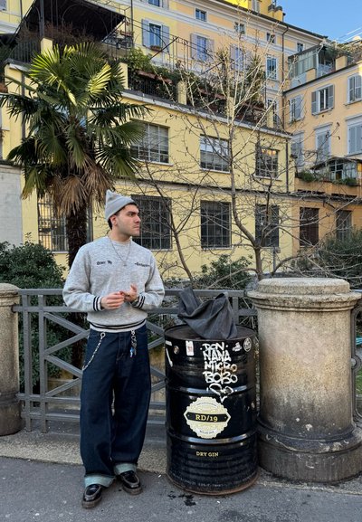 Una persona con un sudadera gris con rayas negras y unos pantalones oscuros holgados está de pie junto a un barril negro cubierto de pegatinas, con una palmera de fondo.