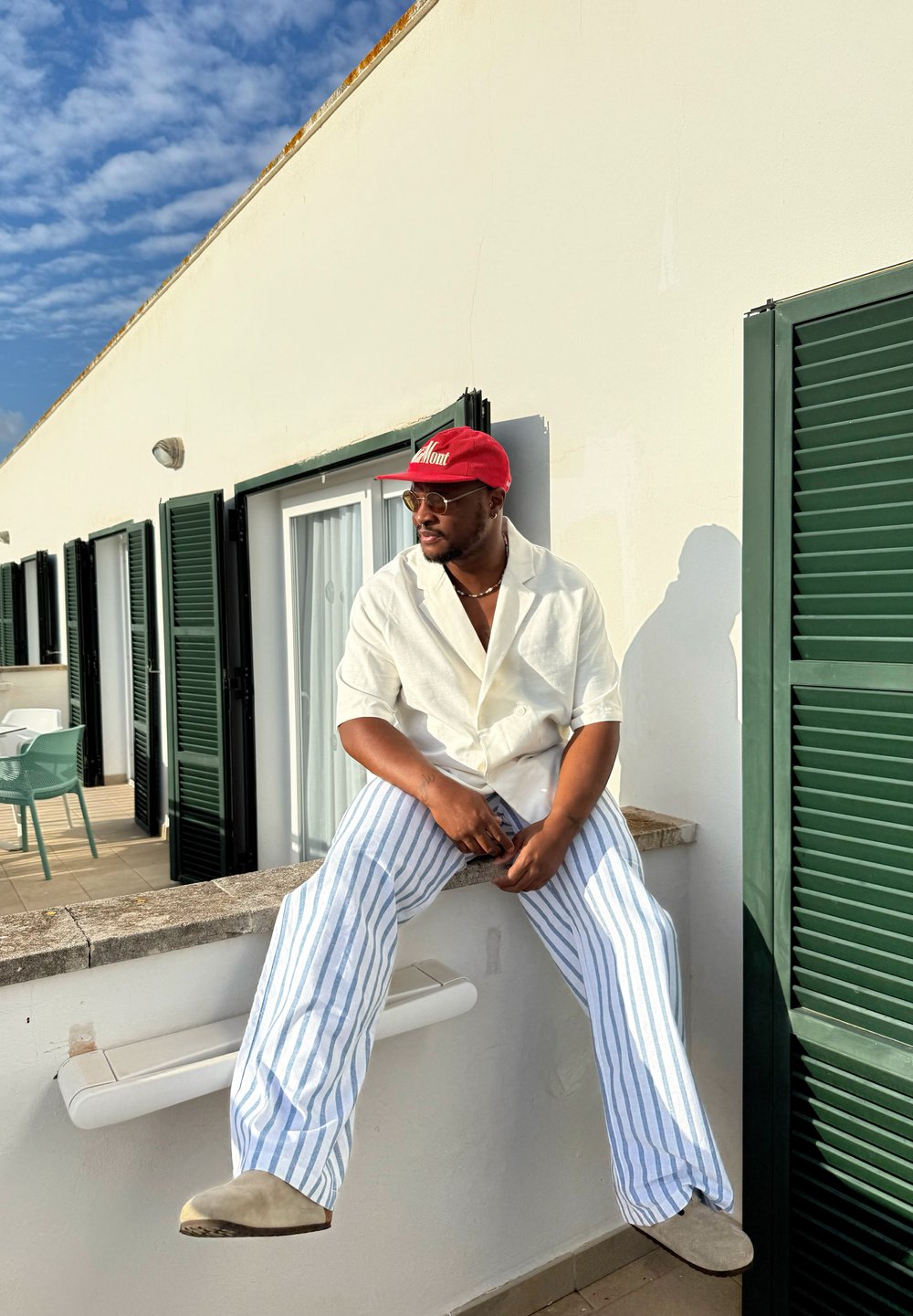 Man wearing a red cap, glasses, a white shirt, and blue-striped trousers sitting on a white balcony wall with green shutters under a partly cloudy sky.