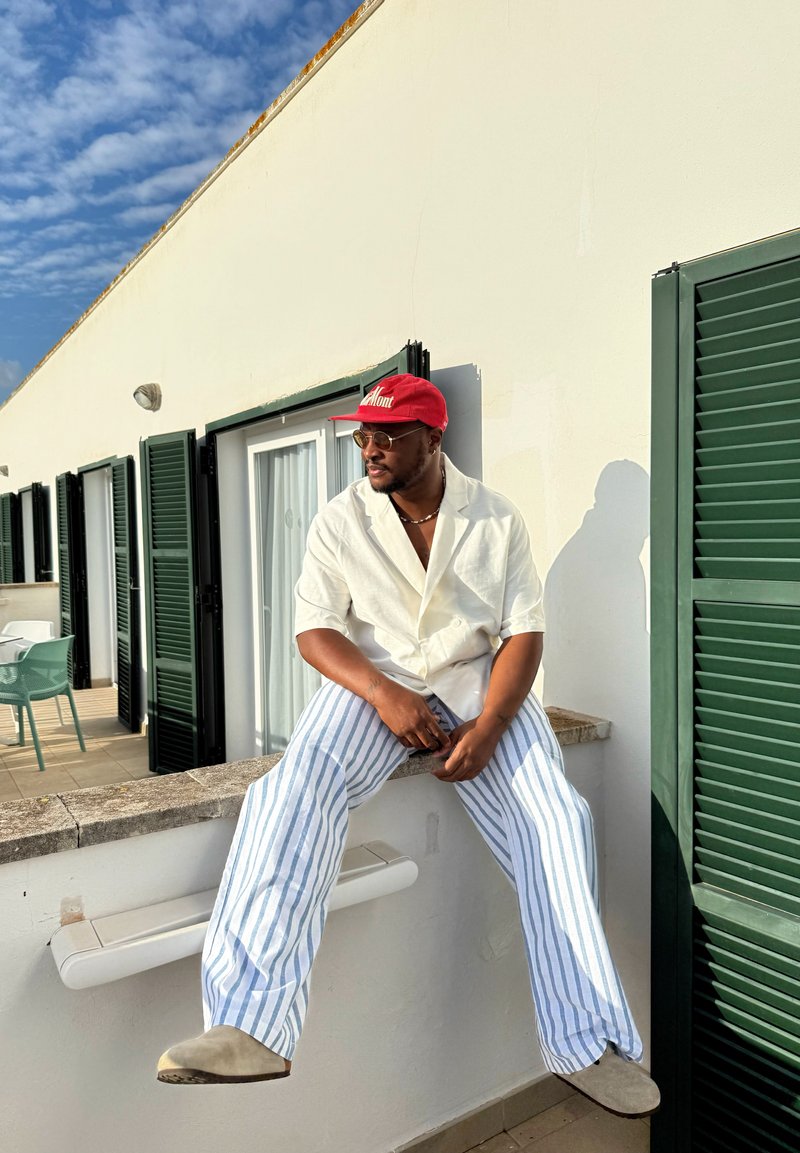 Man wearing a red cap, glasses, a white shirt, and blue-striped trousers sitting on a white balcony wall with green shutters under a partly cloudy sky.