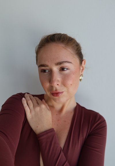 Top largo de color burdeos con un escote en V profundo. Pendientes de aro dorados. Textura suave, piel clara con pecas. Fondo neutro.