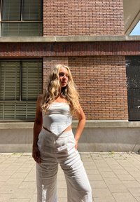 Witte cropped top met spitse zoom, gecombineerd met vloeiende witte linnen broek. Op de achtergrond is een gemêleerde bakstenen muur en ramen met luiken te zien.