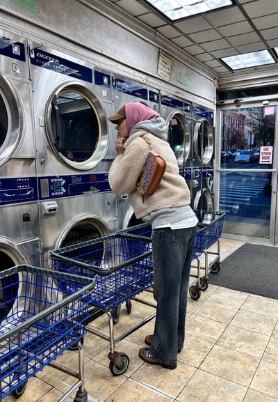 Dos carritos de lavandería azul-gris con ruedas en una lavandería; una persona con un pañuelo rosa en la cabeza y forro polar beige se inclina hacia unas secadoras plateadas.
