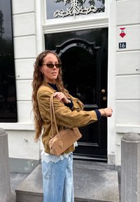 Sac à main en daim marron avec une courte bandoulière, forme rectangulaire, texture lisse ; porté avec un haut en dentelle crème et un jean bleu ample.