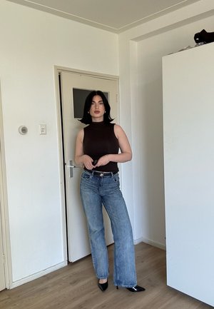 Mujer con cabello negro, top negro sin mangas, jeans azules y tacones negros puntiagudos, de pie en el interior cerca de una puerta cerrada y un armario blanco.