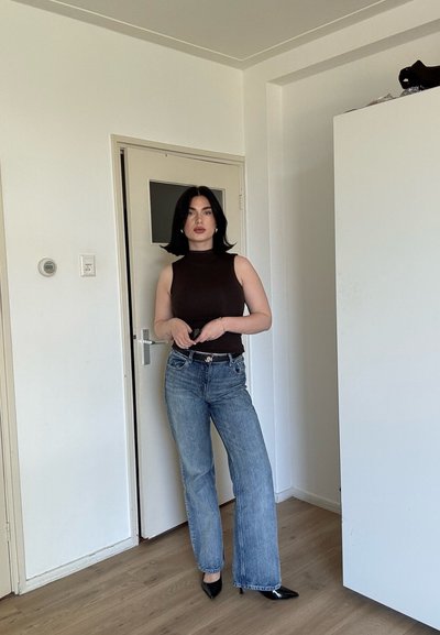 Mujer con cabello negro, top negro sin mangas, jeans azules y tacones negros puntiagudos, de pie en el interior cerca de una puerta cerrada y un armario blanco.