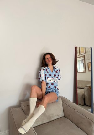 Joven mujer con ropa deportiva azul de lunares y calcetas blancas hasta la rodilla, sentada en un sofá beige, mirando pensativamente hacia arriba cerca de un espejo alto.