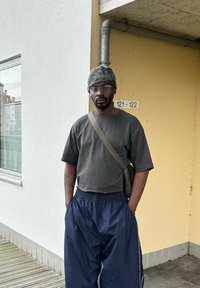 Personne portant une chemise à manches courtes gris foncé et un pantalon ample bleu marine, avec un bonnet à motifs et des lunettes transparentes.