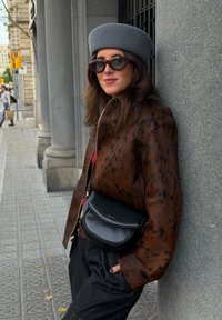 Une femme portant une veste marron à pois, un béret gris et des lunettes de soleil s'appuie contre une colonne en pierre grise sur un trottoir en ville.