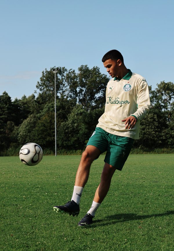 Tenue de sport verte et crème à manches longues, avec un logo et le texte "KidSuper". Un ballon de football noir sur un terrain herbeux, un joueur en mouvement.