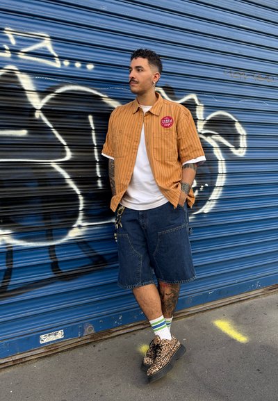 Camisa de manga corta a rayas naranjas con un parche de logo, combinada con shorts de mezclilla oscuros y zapatos de estampado de leopardo. Calcetines blancos con rayas verdes.