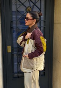 Femme portant une veste matelassée marron, crème et jaune ainsi qu'un pantalon beige, avec des lunettes de soleil et un sac à bandoulière en chaîne, debout près d'une porte en métal noire.