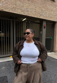 Brązowa zamszowa kurtka na białej prążkowanej koszuli, w połączeniu z beżowymi, wysokim stanem spodniami. Dodatki obejmują okulary przeciwsłoneczne i perłowy naszyjnik.