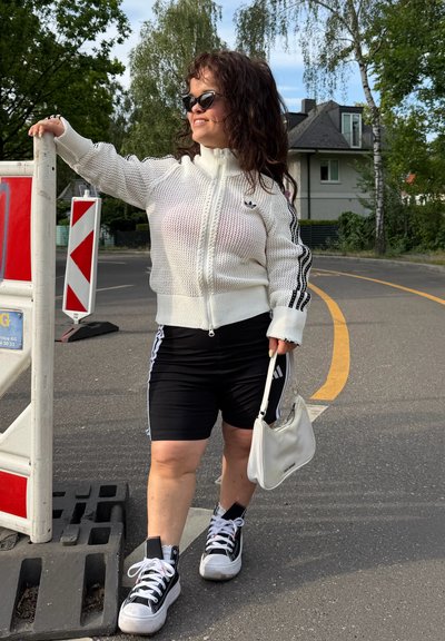 Mujer con chaqueta blanca con cremallera y pantalones cortos negros, sosteniendo una bolsa blanca, parada cerca de un bloqueo en una calle con casas y árboles.