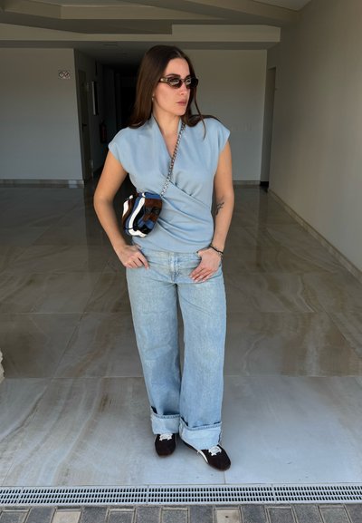 Mujer con top sin mangas azul claro, vaqueros sueltos azul claro, zapatillas oscuras, gafas de sol y bolso bandolera a rayas está de pie en el interior.