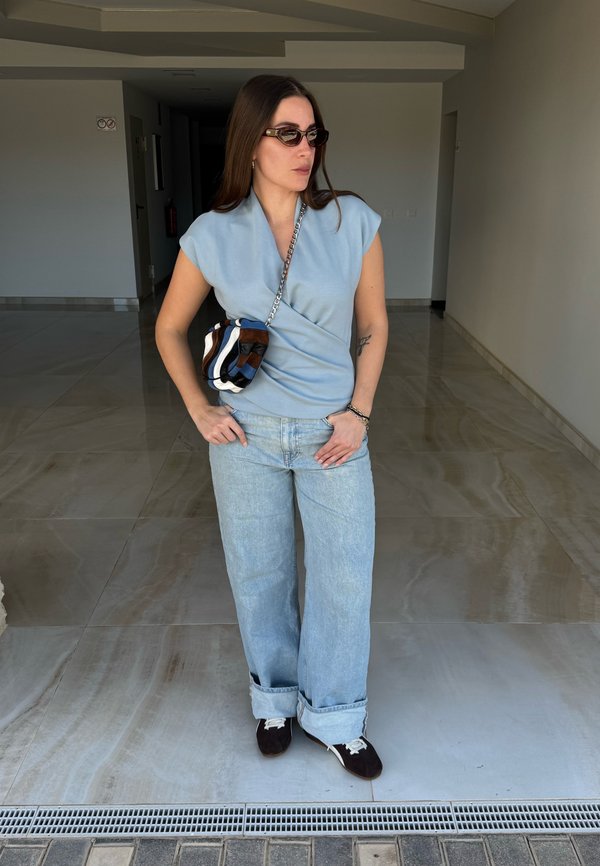 Ženska, ki nosi svetlomodro brezrokavnik, ohlapne svetlomodre kavbojke, temne superge, sončna očala in črto torbico čez ramo, stoji v notranjosti.