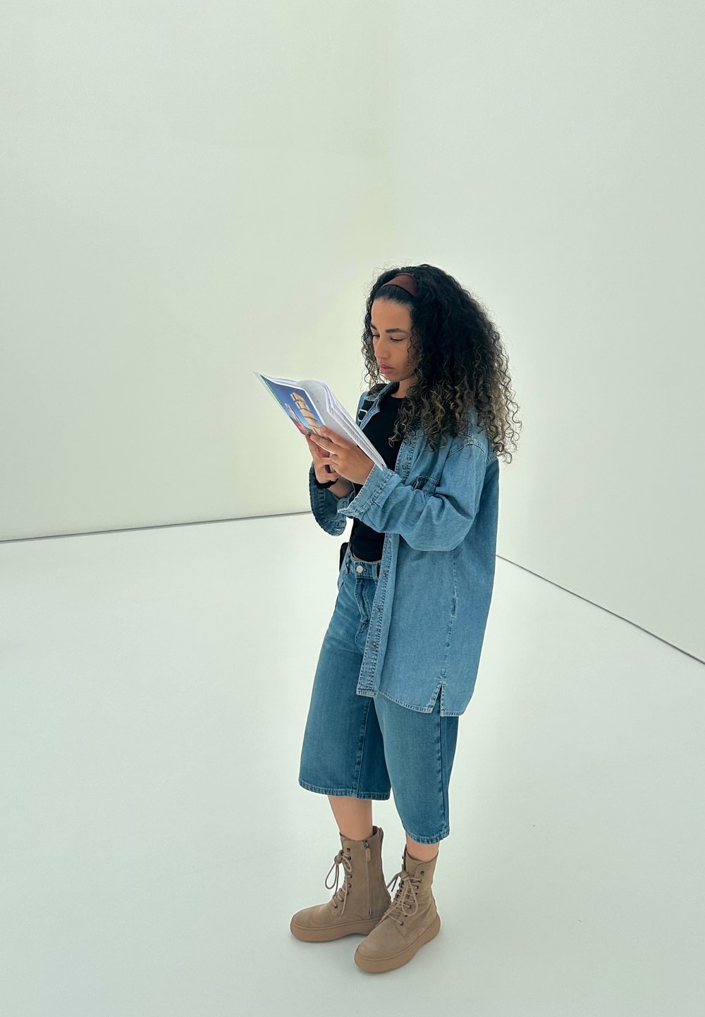 Denimskjorte over svart topp, sammen med bukseshorts i denim som går til knærne og brune snørestøvler. Holder en brosjyre, står i et minimalistisk rom.
