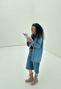 Denimskjorte over svart topp, sammen med bukseshorts i denim som går til knærne og brune snørestøvler. Holder en brosjyre, står i et minimalistisk rom.