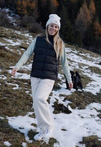 Colete acolchoado preto sem mangas sobre uma blusa de mangas longas azul clara, calças de fleece brancas e sapatilhas brancas, em pé sobre a relva coberta de neve.