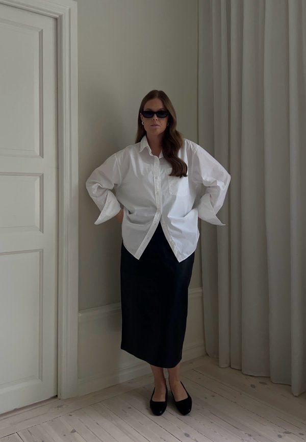 Donna con lunghi capelli castani che indossa occhiali da sole neri, una camicia bianca oversize, una gonna midi nera e scarpe nere, in piedi all'interno.