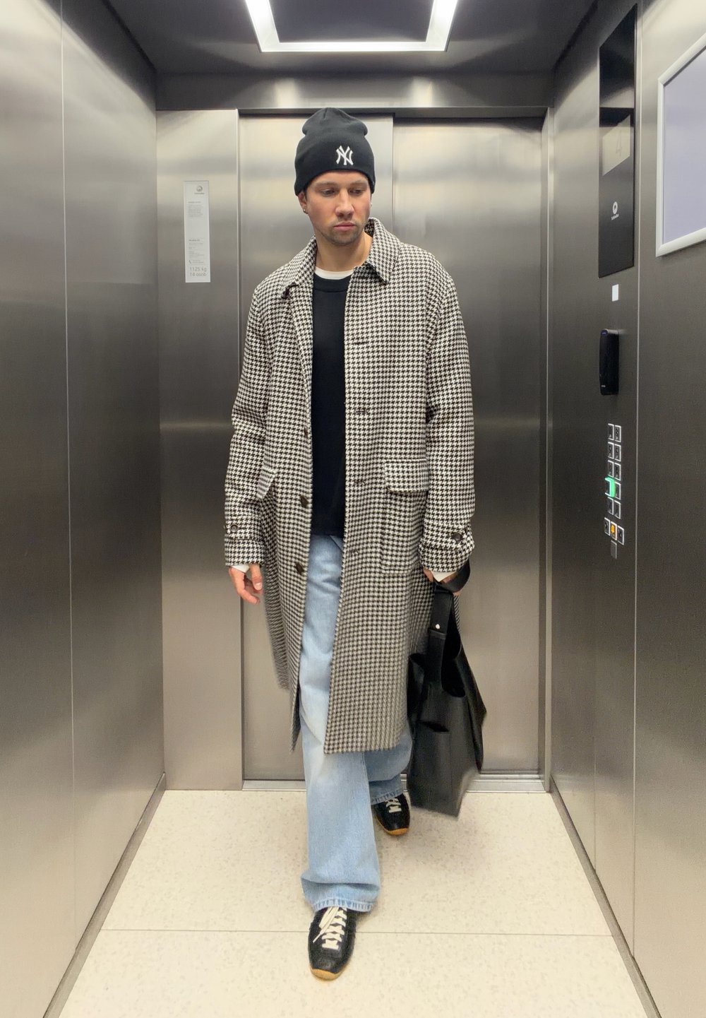 Rutete lang frakk, svart t-skjorte, vide lyseblå jeans, svarte joggesko med brune aksenter, og svart tote bag i en metallisk heis.