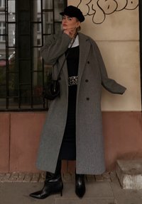 Dlhý sivý vlnený kabát s prehnanými klopami, dvojradovým gombíkovaním a voľným strihom. Nosený cez čierne šaty a v kombinácii s čiernymi koženými čižmami na opätku.