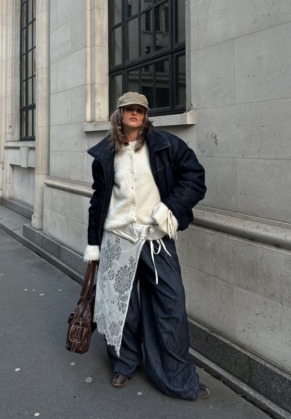 Veste en jean oversize, pull en maille blanc, tablier en dentelle florale, pantalon en jean à jambes larges, casquette beige, sac marron et chaussures marron.