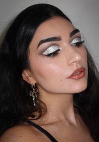 Modell med bølgete svart hår, sølvøyenskygge og glansfulle nakne lepper. Iført en hoop øredobb med detaljer, mot en nøytral bakgrunn.