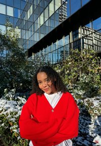 Gilet en tricot rouge avec un décolleté en V profond et de larges manches, porté par-dessus un haut blanc. L'arrière-plan présente de la neige et de la verdure contre un bâtiment en verre.