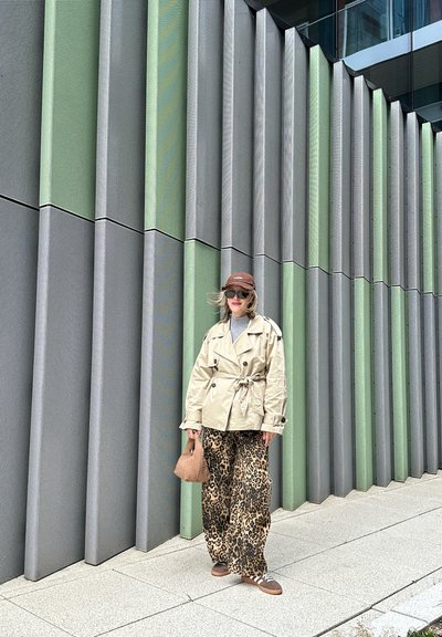 Abrigo trench beige, suéter gris, pantalones de pierna ancha con estampado de leopardo, gorra marrón, gafas de sol y bolso de mimbre, frente a una pared texturizada verde y gris.