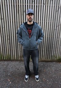 Sweat à capuche gris zippé avec poches avant, t-shirt noir graphique, jean foncé et baskets noires à rayures blanches. Fond en panneaux de bois.