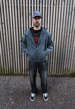 Sudadera con capucha gris con cremallera y bolsillos frontales, camiseta gráfica negra, vaqueros oscuros y deportivas negras con rayas blancas. Fondo de paneles de madera.