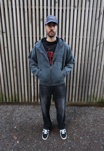 Sudadera con capucha gris con cremallera y bolsillos frontales, camiseta gráfica negra, vaqueros oscuros y deportivas negras con rayas blancas. Fondo de paneles de madera.