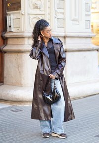 Brauner Trenchcoat aus Kunstleder mit weitem Kragen, kombiniert mit einem schwarzen Top, lockeren blauen Jeans und einer kleinen glänzenden schwarzen Handtasche.