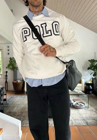 Mann trägt einen weißen "POLO"-Pullover über einem blau gestreiften Hemd, schwarze Hosen, eine Schultertasche und hat sichtbare Tattoos am Hals und an der Hand in geschlossenen Räumen.